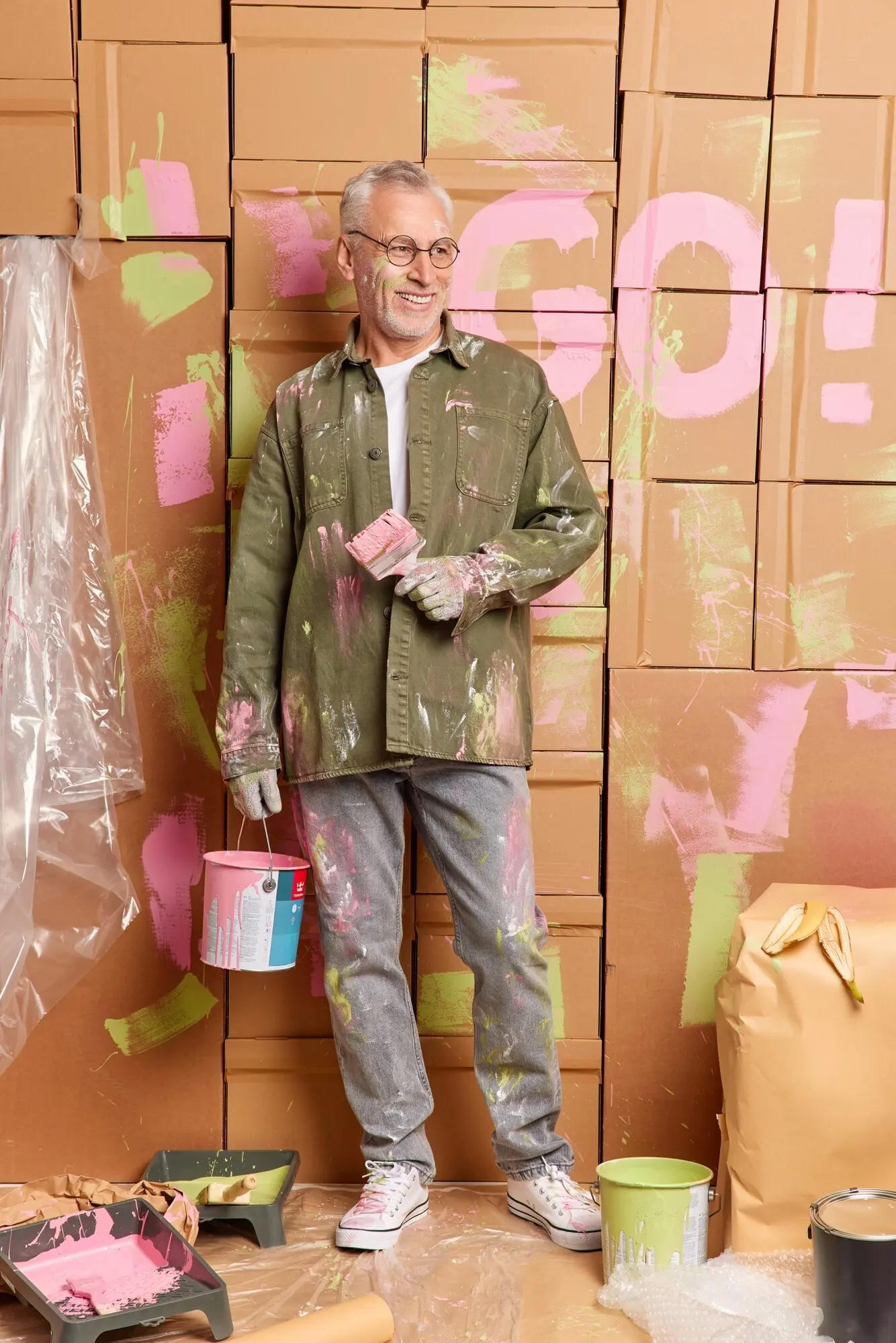 Fröhlicher Senior hält einen Farbeimer und einen Pinsel, führt Reparaturen durch, blickt freudig zur Seite, trägt legere Kleidung und ist zu Hause mit Renovierungsarbeiten beschäftigt. Konzept: Umbau, Hausverbesserung, Instandhaltung.