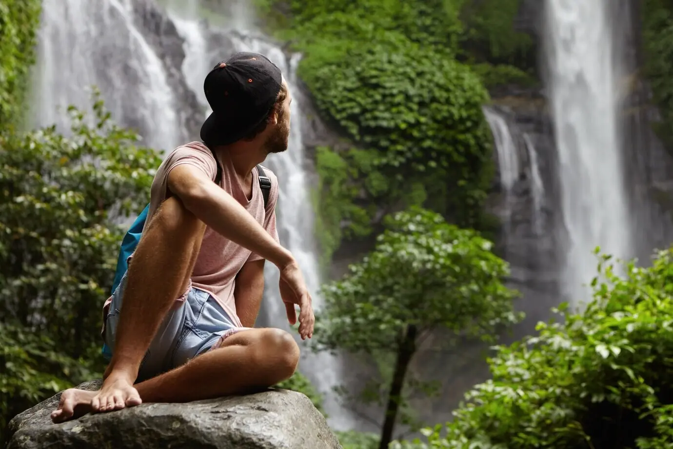 Thema Natur, Tierwelt und Reisen. Ein junger, barfüßiger Wanderer mit Snapback sitzt auf einem großen Felsen und genießt den schönen Ausblick um sich herum. Ein Hipster, der tief im Regenwald entspannt.